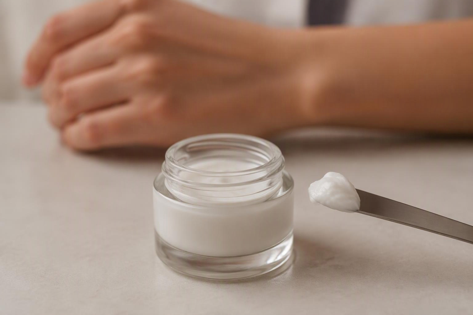 Creme em espátula ao lado de recipiente sem rótulo, com uma mão ao fundo.